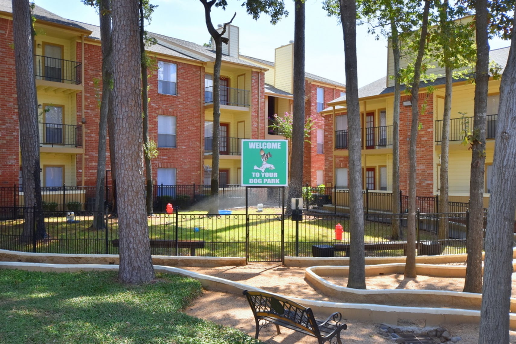 Gated dog park with a small green hill inside, red fire hydrants, and benches to sit. Tall trees and apartment buildings surround the dog park at Deerbrook Apartments.