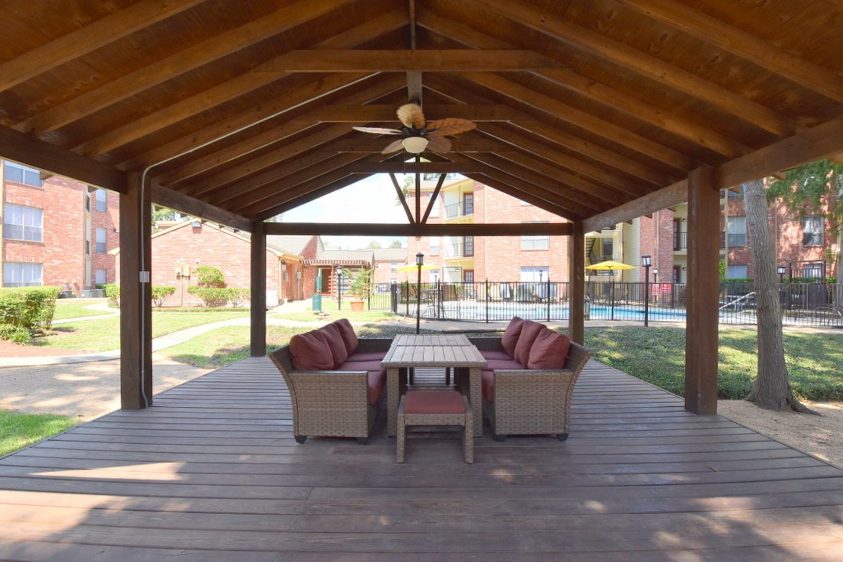 Tall wooden gazebo at Deerbrook Apartments with two wicker couches, two stools, and red cushions. A matching table is between the couches and a  leaf fan above.