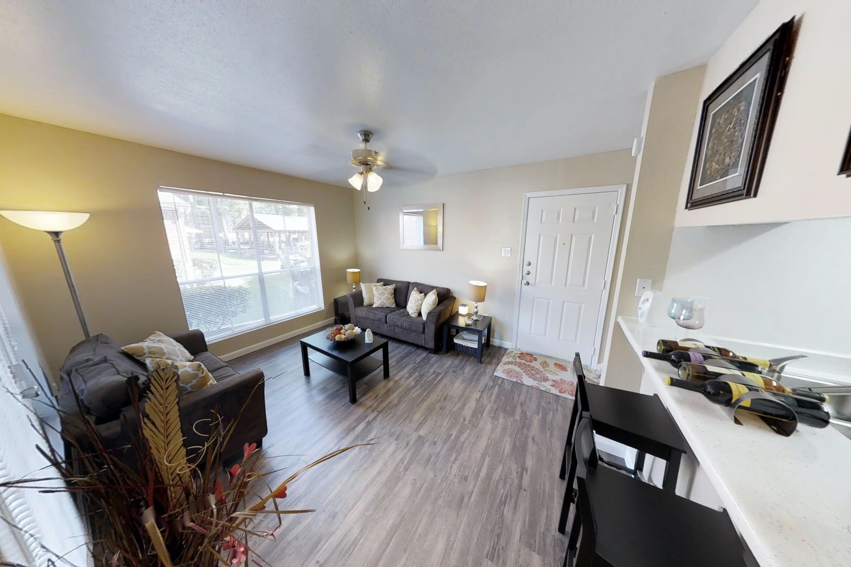 Living space at Deerbrook Apartments with big bright window, brown couches, black side tables and chairs against a counter.