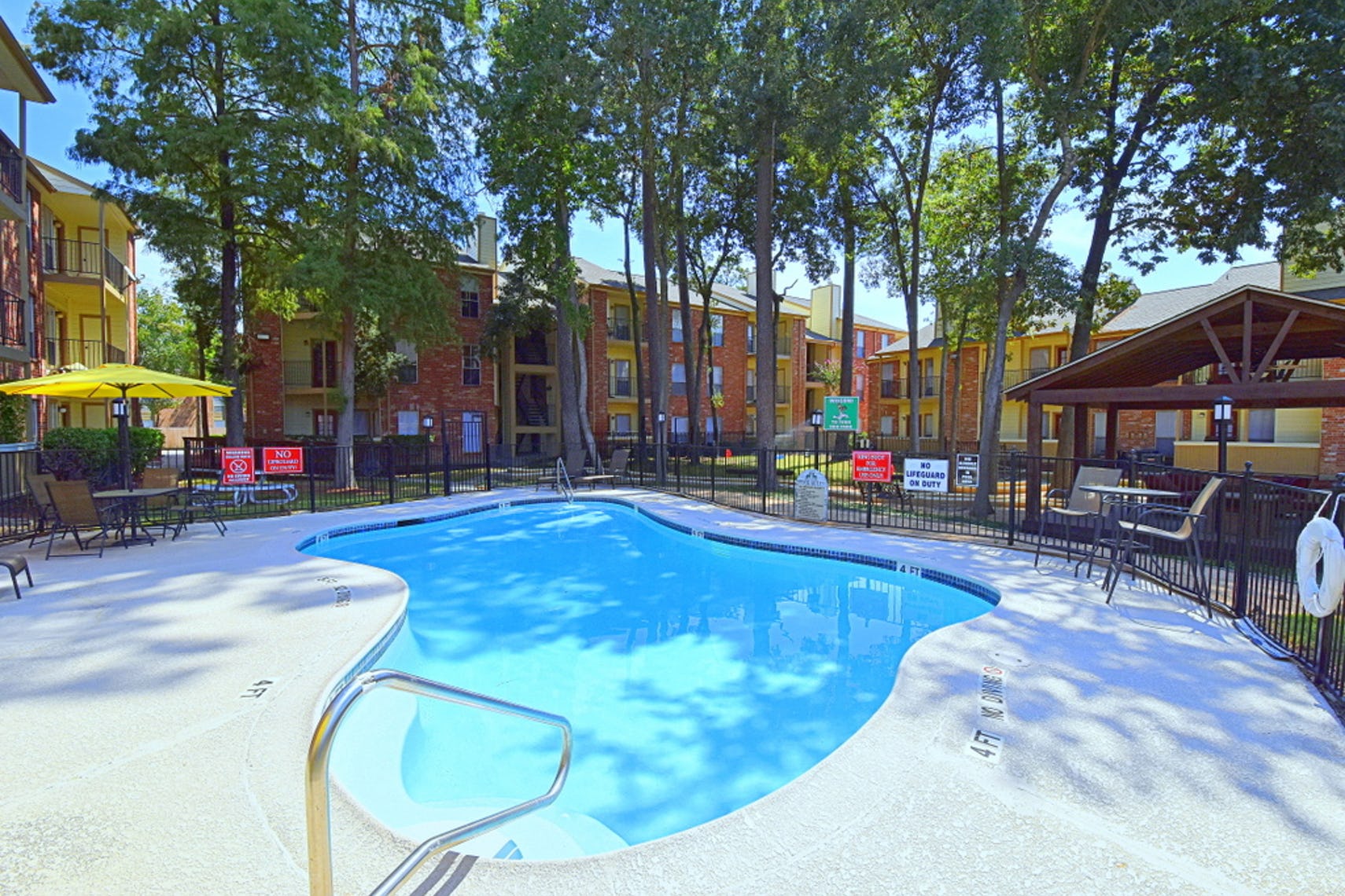 Bright blue pool with outdoor tables, chairs, and a yellow umbrella surrounded by a gate and tall green trees at Deerbrook Apartments.